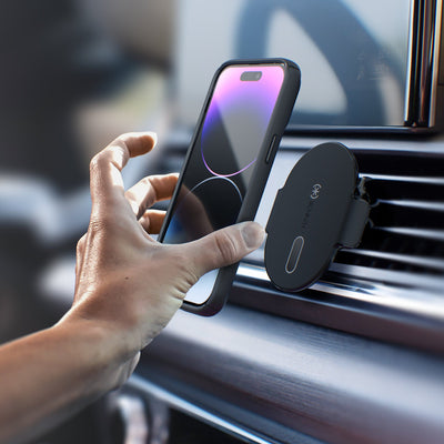 Lifestyle photo of a woman's hand holding a smartphone attaching it to the magnetic car vent mount which is fastened to dashboard vents of a vehicle.#color_black