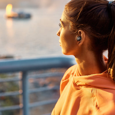 A young woman looks at a sunset over the water with GEMTONES PRO earbuds in her ears. Active noise cancelling silences distractions.#color_black-white