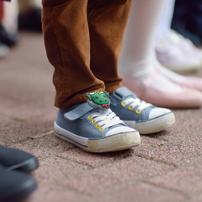 Lifestyle photo of a close-up of a child wearing a shoe in a crowd with Tagimal Blaze, the green dragon, attached to a velcro shoe strap#color_blaze-milo-mittens-and-tink