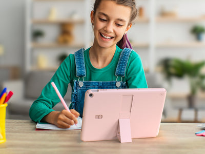 A young girl sits at a table writing in front of a pink Google Pixel Tablet in a StandyShell case by Speck with kickstand out and in view formation