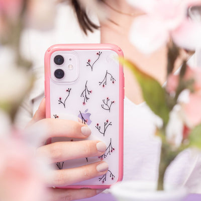 A woman's hand holding Presidio Perfect-Clear + Print: Plum Blossoms Collection case on an iPhone among flowers and blossoms