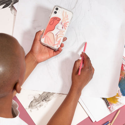 A young woman drawing on paper while holding an iPhone with a Presidio Edition: Blusing Muse Collection case by Speck