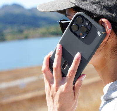 A woman holding an iPhone 15 in a Speck ClickLock grey colored case to her ear with water and hills in the background.